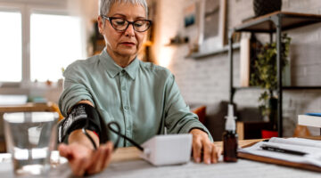 A senior woman sits at the table and measures blood pressure at her home.