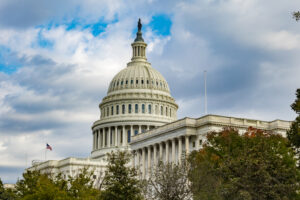 The US Capitol Building in Washington DC, at an angle, over some trees. The beautiful dome stretches up into the blue sky with scattered clouds.