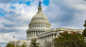 The US Capitol Building in Washington DC, at an angle, over some trees. The beautiful dome stretches up into the blue sky with scattered clouds.
