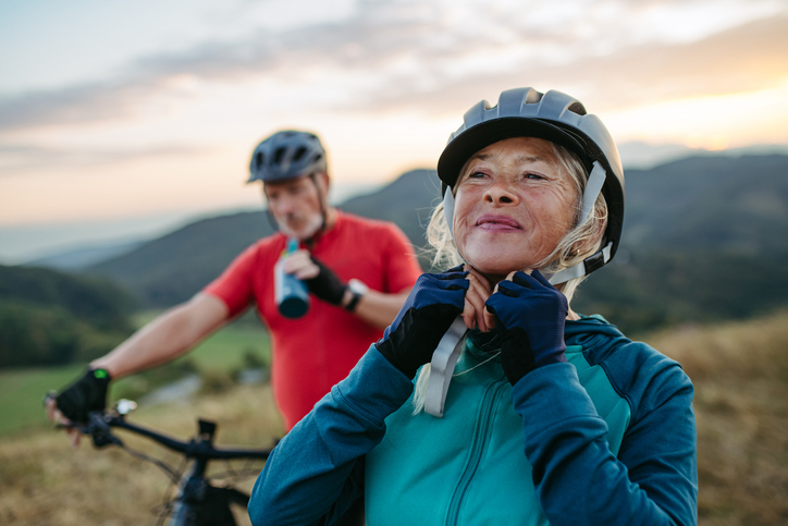 Portrait of beautiful senior couple on bike ride in autumn nature. Taking break, drinking water from sport bottle and fastening helmet.