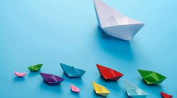Leadership concept using big white paper ship among other colors on a blue background