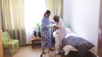 Front view of mixed race female nurse helping senior mixed race female patient to stand with walker at nursing home