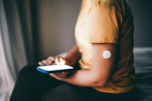 An unrecognizable person using the glucose monitor on the arm, checking the mobile app for more blood sugar level data. The wearable health sensor interacts with a smartphone application, highlighting modern health technology and remote monitoring applications for fitness and wellness.