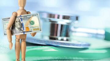 Wooden mannequin holding a US $100 dollar bill on a defocused stethoscope background.