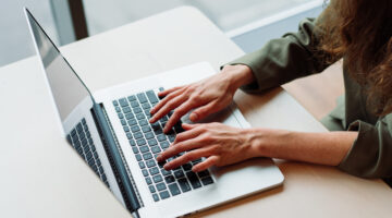 A close-up shot of a professional woman working on a laptop at a modern office desk. Her hands are actively typing on the keyboard, symbolizing productivity, remote work, and digital communication. The setting features a minimalistic design with natural light coming through large windows