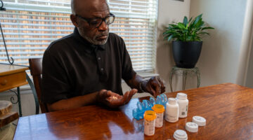 An older African American man with glasses carefully arranges his daily medications in a blue pill organizer. He sits at a wooden table near a window with blinds, methodically sorting prescription bottles and preparing his medication schedule.
