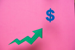 Green upward arrow pointing to a blue dollar sign on a pink background representing increasing profits, financial success, and economic growth