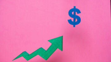 Green upward arrow pointing to a blue dollar sign on a pink background representing increasing profits, financial success, and economic growth