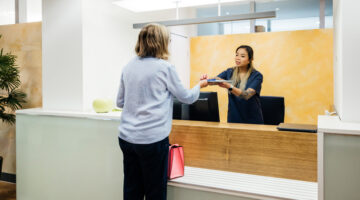 A receptionist in a doctor's office hands paperwork to a patient over the counter. The office is bright and modern, providing a welcoming atmosphere.