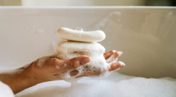 White toilet soap in female hands against the background of a fragrant foam bath. Woman in white terry bathrobe washing her hands with soap. Relaxation and beauty treatments. Natural beauty, daily skincare routine. Moisturizing, cleansing