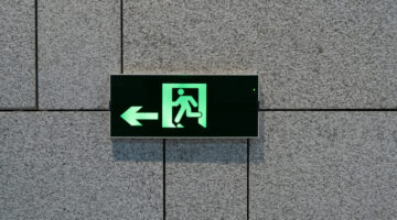 Clear emergency exit sign featuring a green running figure and left arrow, mounted on a textured gray wall, conveying safety and guidance.
