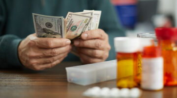 Close-up of a person's hands counting American currency (USD) near several prescription pill bottles and a weekly pill organizer. A strong conceptual image representing high healthcare costs, expensive medication, medical debt, or the affordability of drugs.