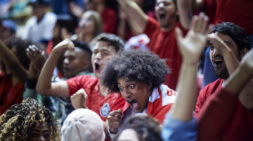 Extremely enthusiastic sports fan with team flag makes face of disbelief and shouts with other fans standing in crowded stadium bleachers after their favorite football team scores a goal in a competitive world sports championship